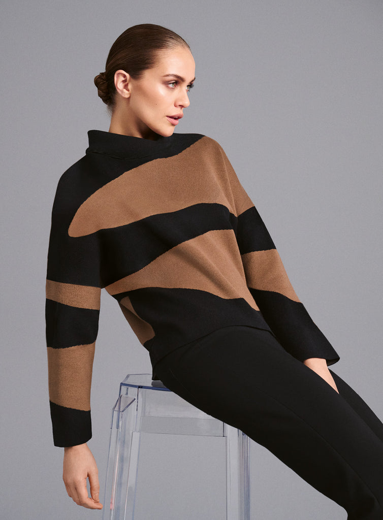 Model trägt weichen Feinstrickpullover mit lockerem Rollkragen in Camel- und Schwarztönen. Breite, geschwungene Streifen sorgen für einen modernen, femininen Look mit grafischem Akzent.
