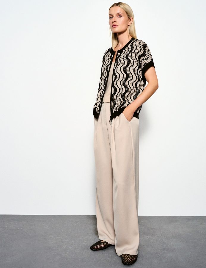 Model trägt einen schwarz-beigen Cardigan mit Zickzackmuster über einem beigefarbenen Top und weiter, fließender beigefarbener Hose; dazu schwarze Loafer mit Cut-outs.