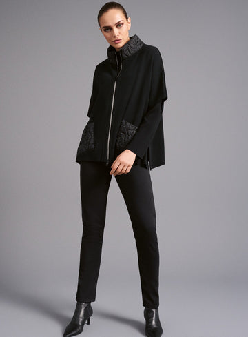 Modell trägt ein komplett schwarzes Outfit bestehend aus einer schmal geschnittenen Hose und einer Jacke im Cape-Stil. Die Jacke besitzt einen Reißverschluss und modische Steppdetails an Kragen und Taschen. Kombiniert mit schwarzen Stiefeletten entsteht ein eleganter und zeitloser Look.