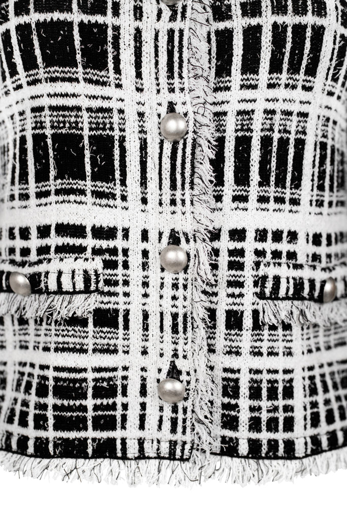 Detailansicht Knopfleiste - Klassische Bouclé-Jacke in weiß mit schwarzem Karomuster, durchgehender Knopfleiste und dekorativen Fransenabschlüssen an Kragen, Saum und Ärmeln. Zwei kleine Pattentaschen auf Hüfthöhe unterstreichen den eleganten Look im Chanel-Stil.