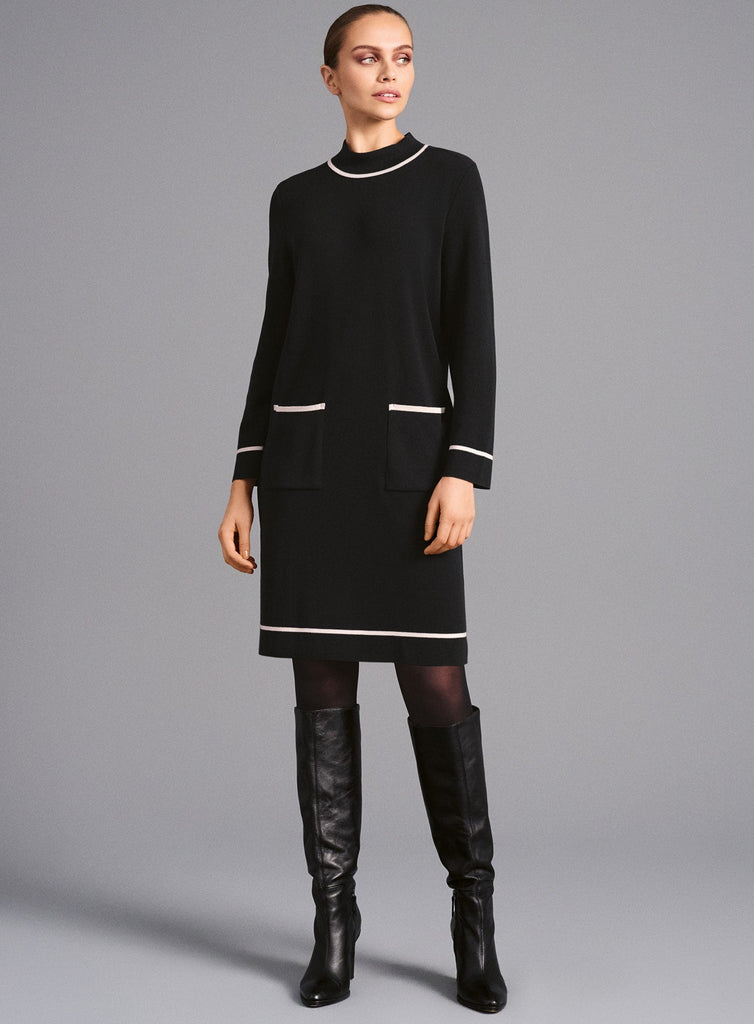 Modell trägt elegantes schwarzes Kleid in knieumspielender Länge mit cremefarbenen Kontraststreifen an Kragen, Ärmeln, Saum und Taschen. Kombiniert mit schwarzen, kniehohen Lederstiefeln entsteht ein klassisch-modernes Outfit.