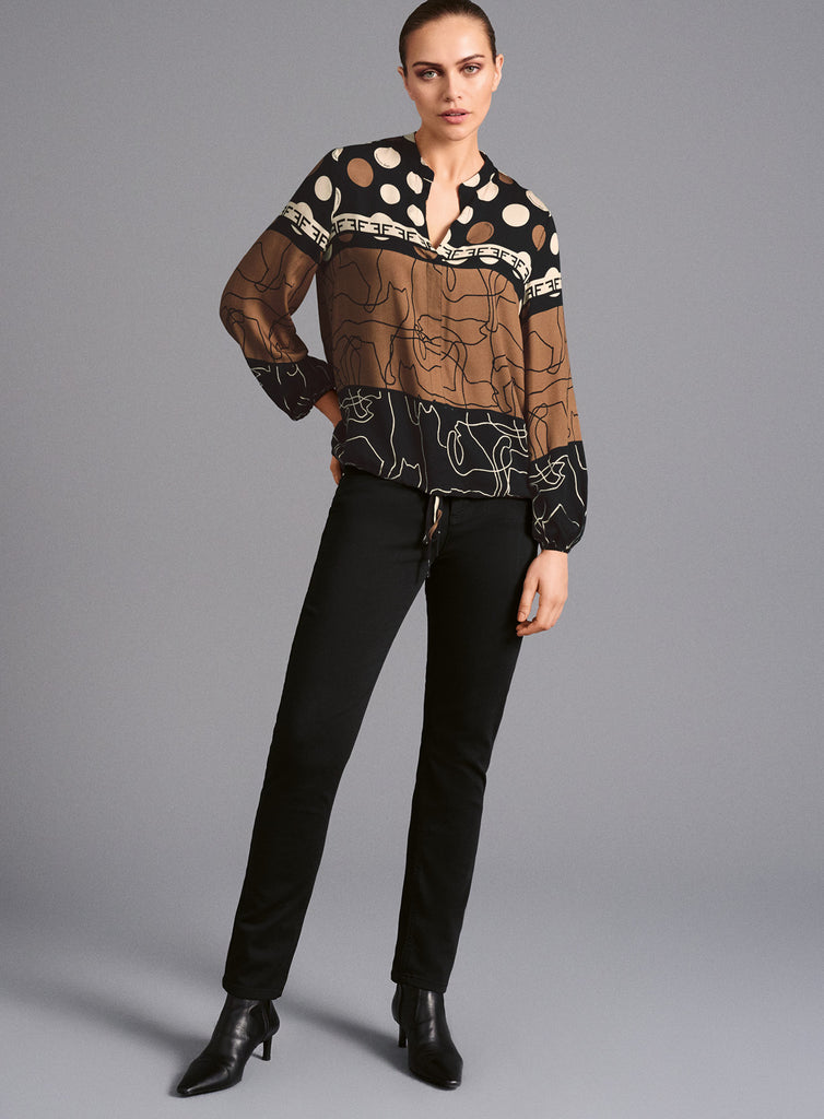 Model trägt aangärmlige Bluse mit vielfältigem Mustermix in Schwarz, Braun und Weiß. Die Bluse kombiniert Punkte, Linienzeichnungen und grafische Elemente, hat einen kleinen Stehkragen, V-Ausschnitt sowie einen Tunnelzug am Saum für variablen Sitz.