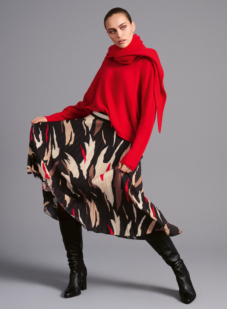 Model trägt plissierten Midirock mit abstraktem Allover-Print in Schwarz, Braun, Weiß und Rot. Der elastische Bund mit sportlichem Streifen sorgt für angenehmen Sitz und modernen Kontrast zum fließenden Stoff. Dazu einen roten Pullover und einen roten Schal.