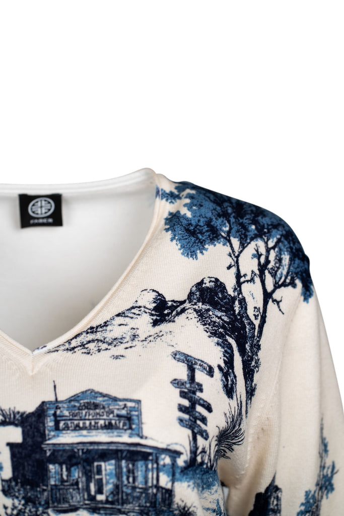Detaillansicht des Ausschnitts des cremefarbenen Pullovers mit V-Ausschnitt und einem detaillierten, blauen Landschaftsmotiv. Das Design zeigt stilisierte Natur- und Gebäudemotive und verleiht dem Pullover einen modischen, eleganten Charakter.
